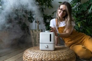 Happy woman gardener sitting on wooden floor, using air humidifier in home garden. Plant care.